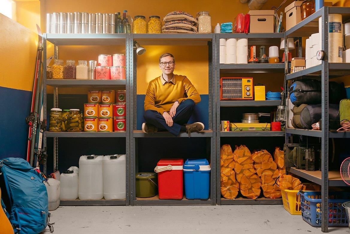 et lager med ting til egenberedskap. En person sitter også i rommet, og illustrerer en beredskapsvenn. - Klikk for stort bilde